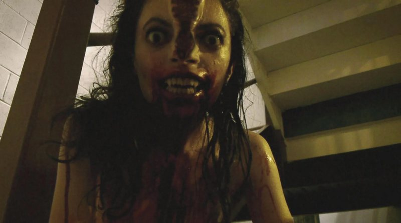 Screenshot do filme de horror V/H/S (2012). A imagem mostra uma mulher em primeiro plano, com o rosto coberto de sangue, cabelo molhado e desgrenhado. Seus olhos estão arregalados e fixos na câmera, e sua boca aparece aberta, revelando dentes também sujos de sangue. O ângulo baixo e próximo reforça a sensação de ameaça, típica do gênero found footage. Ao fundo, é possível ver parte de uma escada, mas o destaque total da cena é a figura feminina em um momento de terror visceral.