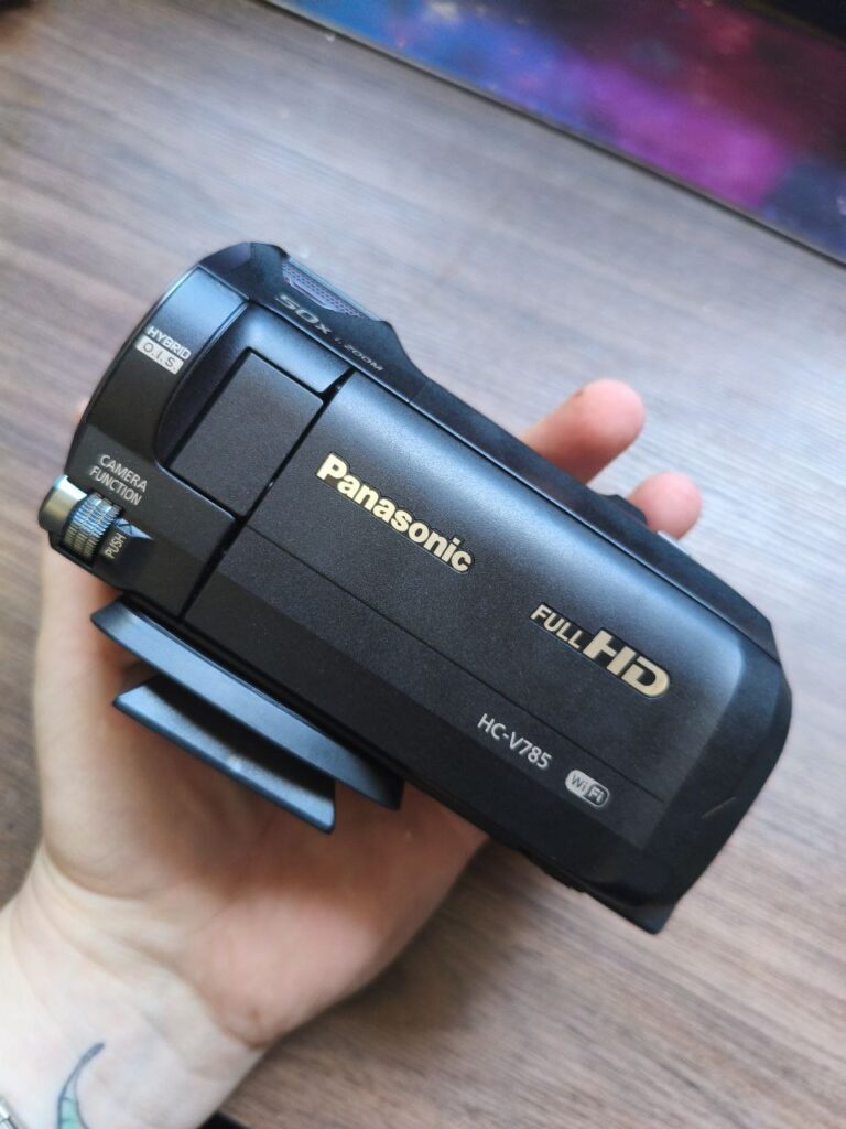 Filmadora Panasonic Full HD preta, modelo HC-V785, sendo segurada na mão. O equipamento está fechado, com destaque para a inscrição “Panasonic Full HD” em dourado e a lente embutida. O acabamento é fosco e compacto, com botões laterais para funções da câmera.