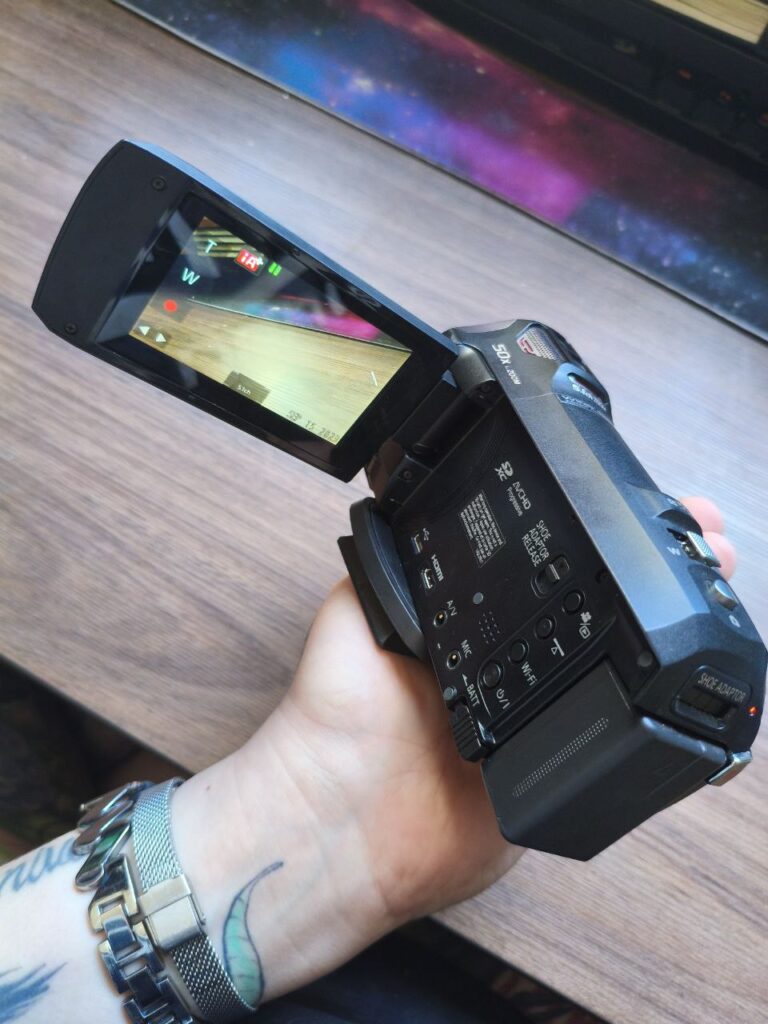 Filmadora Panasonic preta sendo segurada na mão, com a tela lateral aberta. No visor, aparece a gravação de uma superfície de madeira clara. A lateral do equipamento mostra entradas de áudio e conexões, com detalhes de botões de controle visíveis.