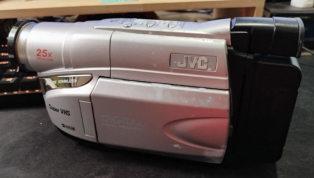 Filmadora JVC prateada posicionada sobre uma mesa escura. O equipamento está fechado, exibindo a inscrição "Super VHS", "25x Optical Zoom" em vermelho na lente e "Digital Signal Processing" na lateral. A textura mostra sinais de uso, com algumas manchas na superfície.