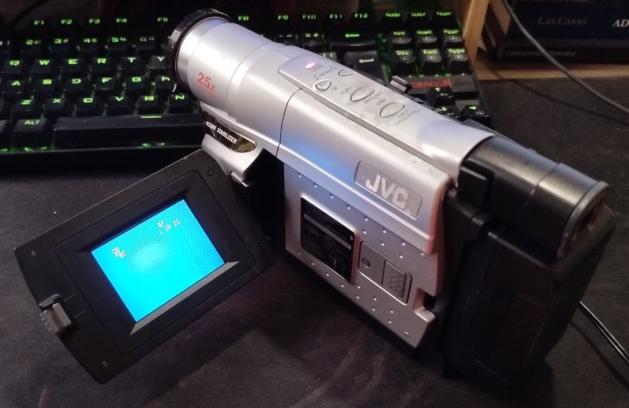 Filmadora JVC prateada em formato horizontal, com lente à esquerda e visor LCD aberto mostrando uma tela azul. O equipamento está sendo segurado por uma mão sobre uma mesa escura, com teclado iluminado em verde no fundo.