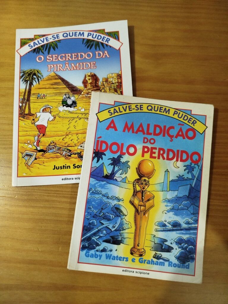 Dois livros da coleção “Salve-se Quem Puder”: “A Maldição do Ídolo Perdido” e “O Segredo da Pirâmide”, da editora Scipione, sobre uma mesa de madeira.