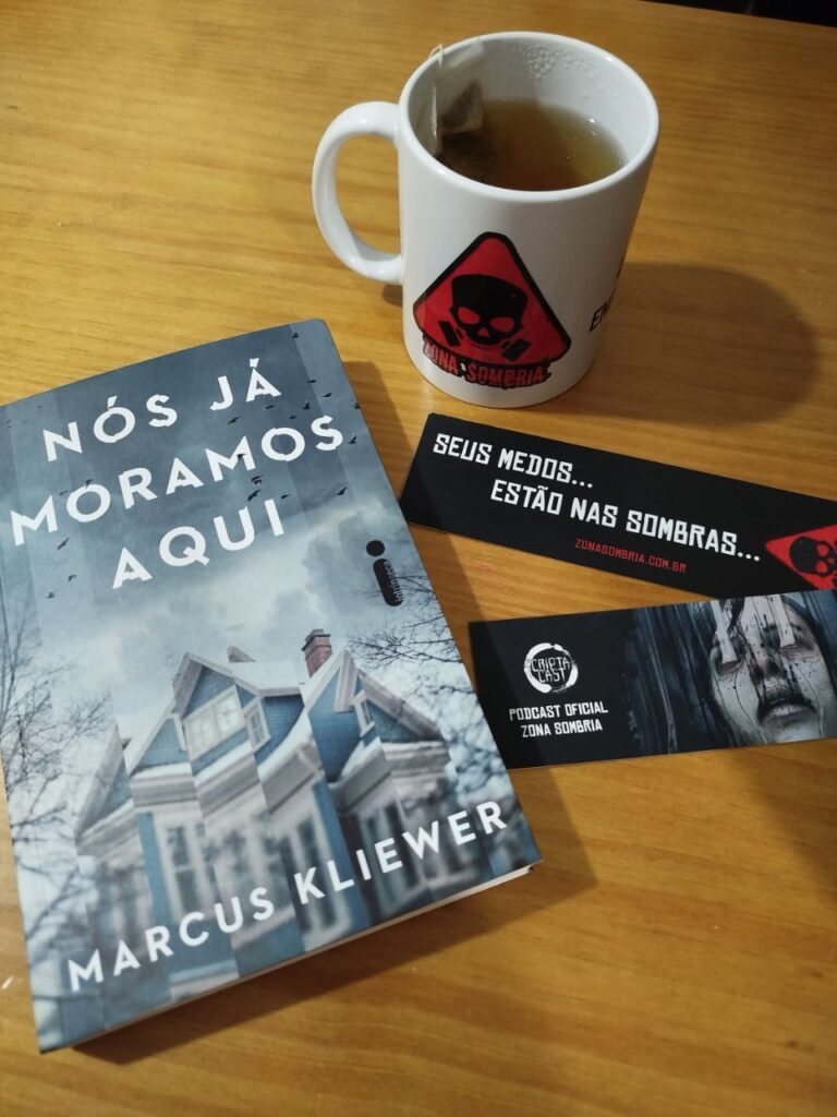 Livro Nós Já Moramos Aqui, de Marcus Kliewer, sobre uma mesa de madeira. Ao lado, há uma caneca branca com o logo vermelho do Zona Sombria e dois marcadores de página pretos — um com a frase “Seus medos... estão nas sombras...” e o outro com o rosto assustador de uma mulher e o logo do CriptaCast.